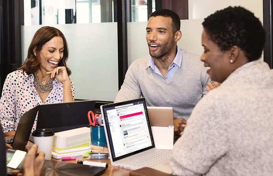 Dans un bureau moderne, trois personnes sourient et discutent autour d'une table avec des PC utilisant Office 2019 Pro Plus.