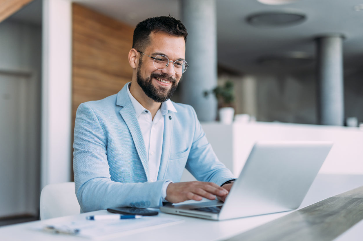 Homme souriant avec barbe et lunettes en blazer bleu clair, assis à un bureau moderne, utilise un ordinateur portable ; stylos, papiers et smartphone présents.