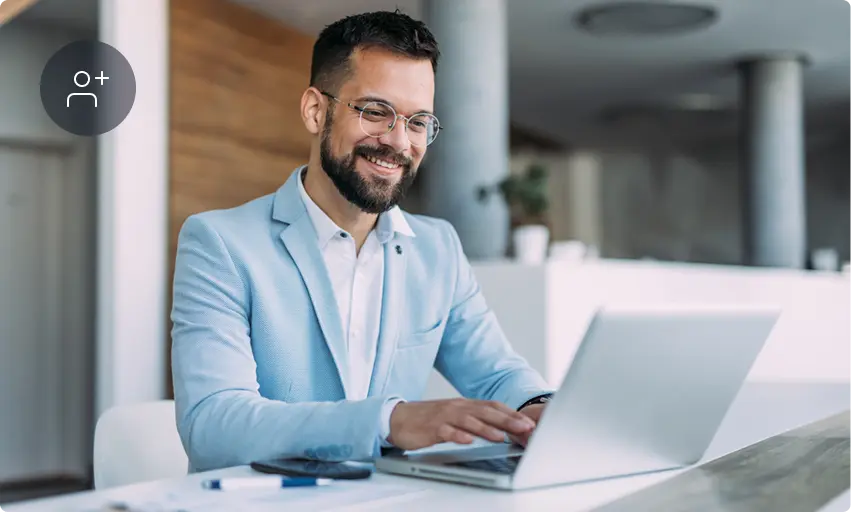 Homme souriant à lunettes et barbe, en blazer bleu clair, travaille sur ordinateur portable dans un bureau moderne lumineux ; icône ajout contact.