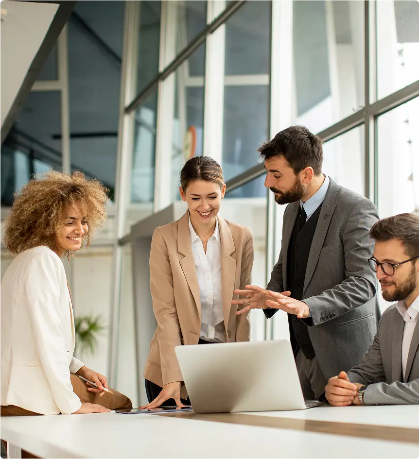 Quatre professionnels autour d'une table moderne discutent et sourient devant un ordinateur portable, éclairés par de grandes fenêtres.