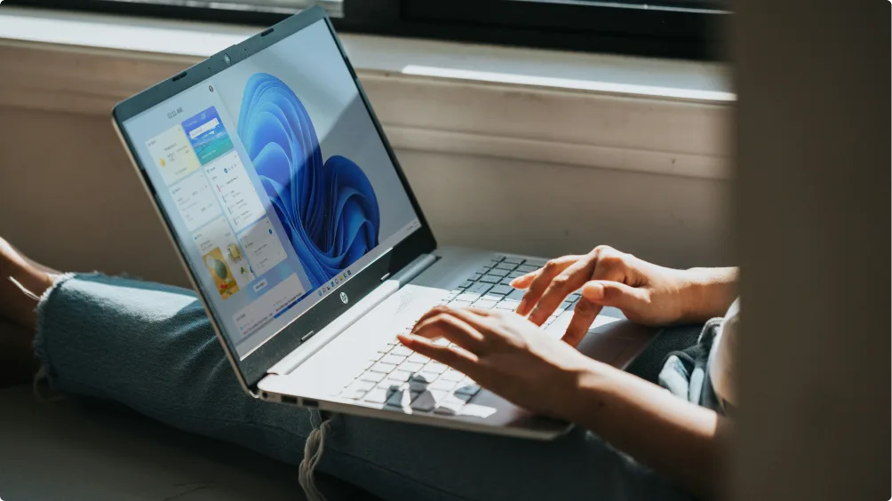 Personne assise au sol, tapant sur un ordinateur portable HP blanc affichant l'accueil Windows 11, baignée par la lumière du soleil.