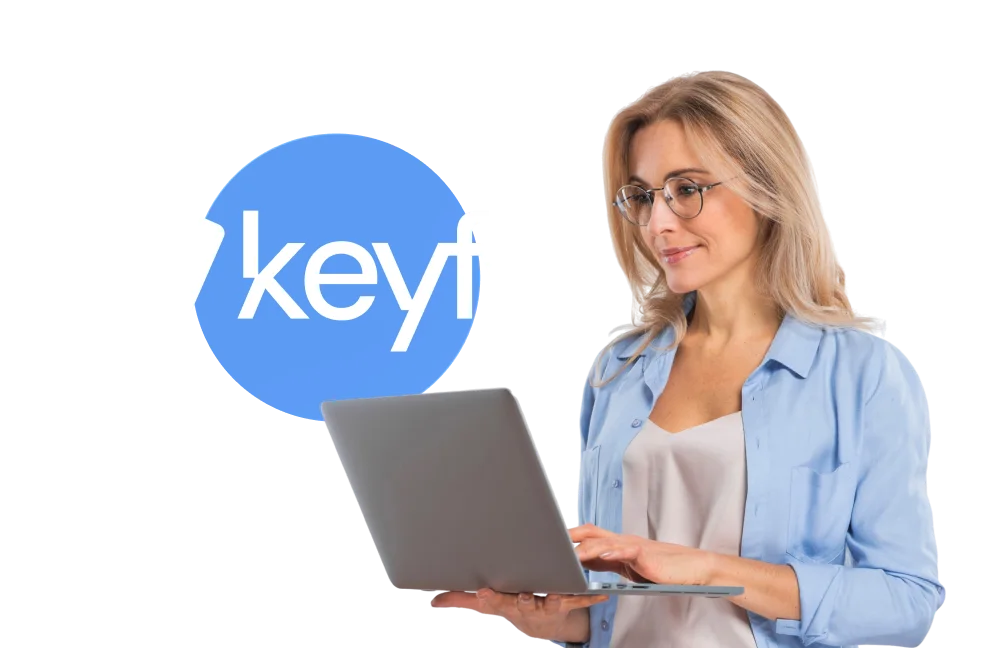 Une femme blonde à lunettes, en chemise bleu clair, consulte un ordinateur portable devant le logo keyfy sur fond bleu.