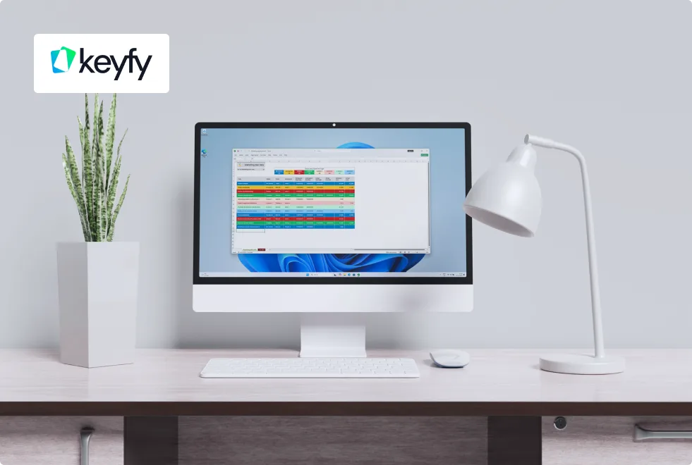 Bureau moderne avec ordinateur affichant un tableau coloré, lampe blanche, clavier et souris blancs, plante verte et logo Keyfy au mur.