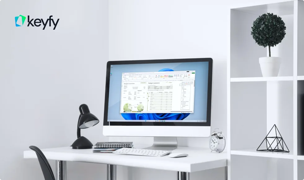 Bureau blanc moderne avec ordinateur affichant un tableur, lampe de bureau, carnet, réveil et étagère décorative. Logo Keyfy au mur.