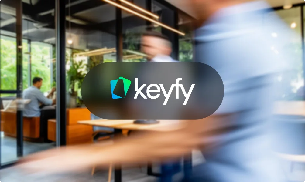 Des personnes floues marchent dans un bureau moderne à parois vitrées, deux hommes discutent. Logo Keyfy bleu et vert au premier plan.